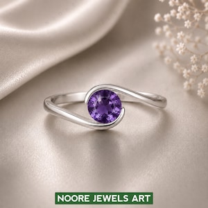 Könnte beinhalten: Ein silberner Ring mit einem runden, tiefvioletten Edelstein, der in einem wirbelnden Design gefasst ist. Der Ring ist vor einem weichen, neutralen Hintergrund platziert. Der Text "NOORE JEWELS ART" befindet sich am unteren Bildrand.