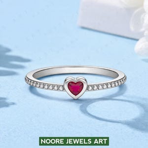 Könnte beinhalten: Ein silberner Ring mit einem herzförmigen, rubinroten Edelstein in der Mitte. Das Band ist mit kleinen, runden Verzierungen versehen. Der Ring befindet sich vor einem hellblauen Hintergrund. Der Text "NOORE JEWELS ART" steht unten.