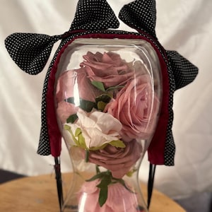 Op de afbeelding: Een heldere glazen vaas gevuld met roze en witte rozen, versierd met een zwarte haarband met witte stippen en rode rand. De vaas heeft een zandloper vorm, waardoor een decoratief bloemstuk ontstaat.