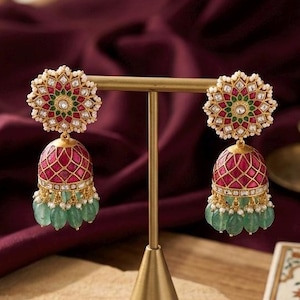 May include: A pair of gold-tone jhumka earrings featuring a circular stud with red, green, and white stones. The bell-shaped drop is decorated with a red grid pattern and finished with a fringe of light green teardrop-shaped beads and small pearls.
