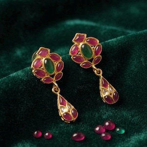 May include: A pair of gold-toned drop earrings featuring a central oval green stone surrounded by six small oval pink stones, with a dangling teardrop-shaped pendant containing a pink stone. Several loose pink and green stones lie nearby.