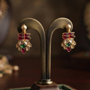 May include: A pair of gold-toned drop earrings featuring a central green stone surrounded by red petal-shaped accents and a small white pearl at the top. The earrings are displayed on a dark metal stand with a wooden base containing loose stones.