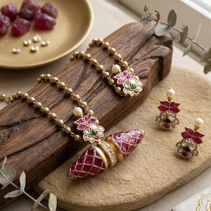 Op de afbeelding: Een goudkleurige ketting en bijpassende oorbellen met robijnrode accenten en een geometrisch patroon. De ketting heeft een centrale hanger met een ruitvormig patroon. De oorbellen en de ketting hebben een bloemenpatroon met parels en groene en witte accenten.