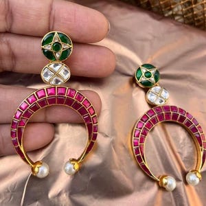 Op de afbeelding: Goudkleurige oorbellen met een halvemaanvormig ontwerp, bezet met rode stenen. De oorbellen hebben een groen en wit bovenstuk en een parel aan de onderkant. De oorbellen worden gepresenteerd tegen een gouden en roze achtergrond.