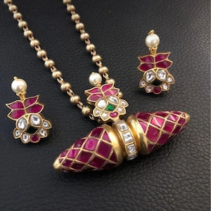 Puede incluir: Un conjunto de joyas de tono dorado que incluye un collar, un colgante y pendientes. El conjunto incluye un collar con una cadena de cuentas, un colgante con un diseño de diamantes de color rubí y pendientes a juego con detalles de perlas. Las joyas están sobre un fondo negro.