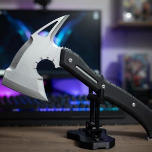 Könnte beinhalten: Eine silberne und schwarze taktische Tomahawk-Axt mit gezahnter Kante und kreisförmigem Ausschnitt. Die Axt hat einen schwarzen Griff und wird auf einem schwarzen Ständer ausgestellt. Der Hintergrund umfasst einen Computermonitor und eine Tastatur.
