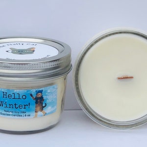 Pode incluir: Duas velas em frascos de vidro com tampas prateadas. Uma tem um rótulo que diz "Hello Winter!" com um gato de desenho animado com roupas de inverno. A outra mostra o pavio de madeira. Cada vela contém 170 g de cera de soja natural.