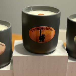 May include: Three gray ceramic candles with wooden wicks. Each candle has a label with a cat-themed design. One says "Meowy Christmas", another "Catmosphere", and the third "Happy Mew Year". The candles are on a white surface.