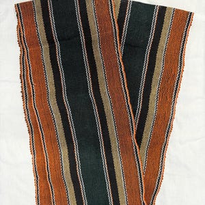Può includere: Tessuto a trama con strisce verticali nei toni dell'arancione, nero, verde, beige e bianco. Il tessuto sembra essere piegato su una superficie bianca. I bordi del tessuto presentano un dettaglio sfrangiato.