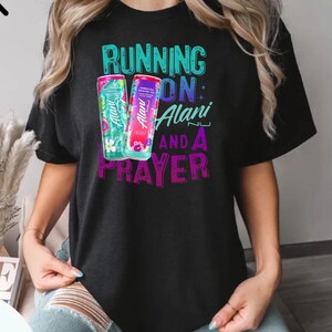 A person wearing a black t-shirt with a graphic design featuring a can of energy drink and the text "RUNNING ON ALANI AND A PRAYER".