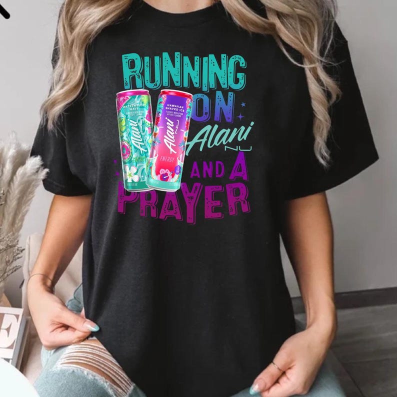 A person wearing a black t-shirt with a graphic design featuring a can of energy drink and the text "RUNNING ON ALANI AND A PRAYER".
