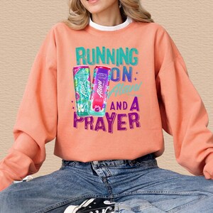 A woman wearing a peach-colored sweatshirt with a graphic design featuring a pair of cans and the text "RUNNING ON ALAN AND A PRAYER".