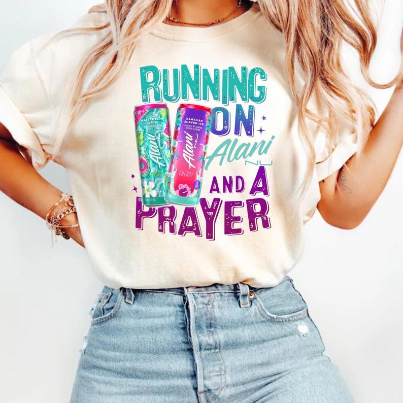 A woman wearing a white t-shirt with a graphic design featuring a bottle of Alani energy drink and the text "Running on Prayer".