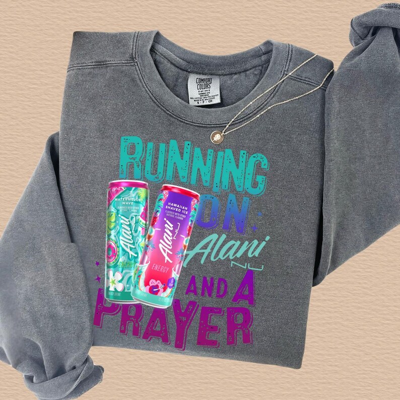 A gray sweatshirt with a graphic design featuring a bottle of Alani energy drink and the text "RUNNING ON ALANI AND A PRAYER".