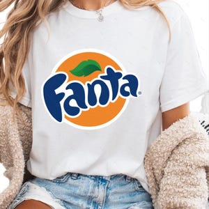 May include: A white short-sleeved t-shirt featuring a large, centered graphic of the blue Fanta logo set against a bright orange circular background with a small green leaf accent above the text. The shirt has a classic crew neckline design.