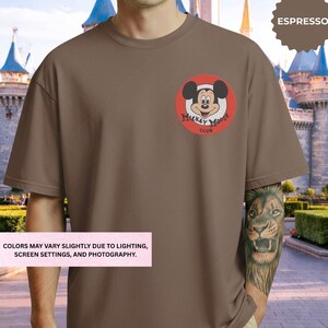 A man wearing a brown t-shirt with a Mickey Mouse logo on it, standing in front of a castle-like structure.