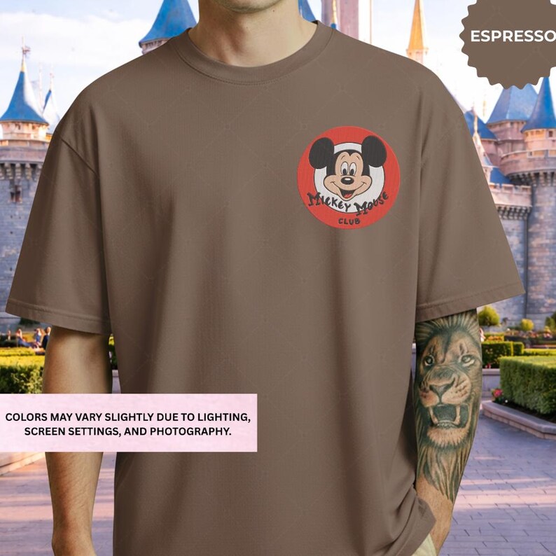 A man wearing a brown t-shirt with a Mickey Mouse logo on it, standing in front of a castle-like structure.
