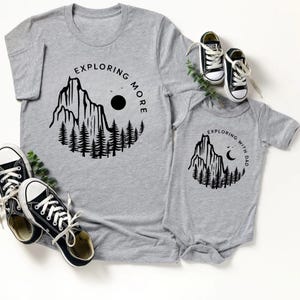 May include: Gray t-shirt and onesie set with black graphic mountain and tree design. The t-shirt says "Exploring More" and the onesie says "Exploring with Dad". Black and white sneakers are also in the image.
