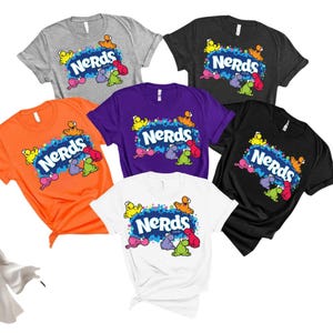 May include: A collection of six short-sleeve t-shirts in gray, black, orange, purple, and white. Each shirt features the blue Nerds candy logo surrounded by colorful, small, cartoon-like character shapes on a background of blue and purple dots.