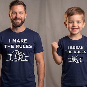 May include: Two navy blue t-shirts with white text and graphic fist bump design. One shirt reads "I MAKE THE RULES" and the other reads "I BREAK THE RULES". The shirts are short-sleeved.