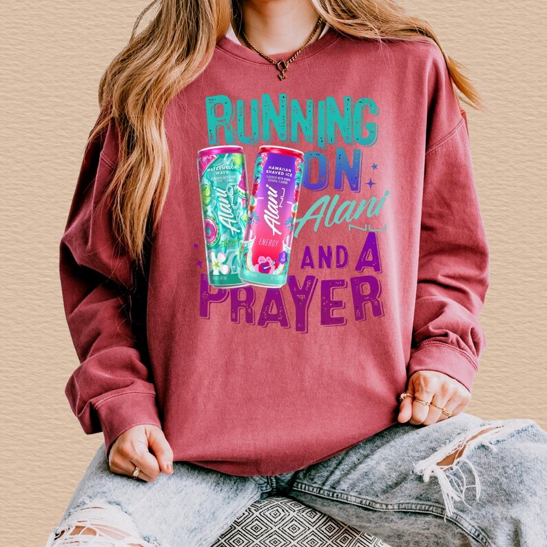 A person wearing a red sweatshirt with a graphic design featuring a can of energy drink and the text "RUNNING ON ALABI" and "AND A PRAYER".