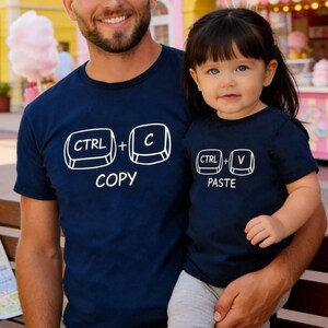 May include: Navy blue t-shirts with white text. One shirt says "CTRL + C COPY" and the other says "CTRL + V PASTE". The shirts are worn by a parent and child, suggesting a matching set.