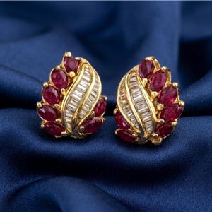 Könnte beinhalten: Ein Paar goldfarbene Ohrringe mit oval geschliffenen Rubin-Edelsteinen und Baguette-Diamanten. Die Ohrringe haben ein blattähnliches Design, wobei die Rubine die äußeren Ränder und die Diamanten die Mitte bilden. Auf blauem Hintergrund.