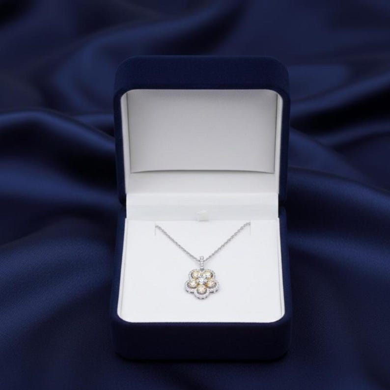 Puede incluir: Un collar de plata con un colgante en forma de flor se muestra en una caja de joyer&iacute;a de terciopelo azul oscuro. El colgante presenta un grupo de peque&ntilde;as piedras preciosas redondas rodeadas por un halo de piedras m&aacute;s peque&ntilde;as. La caja est&aacute; abierta.