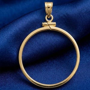 May include: A gold-coloured pendant with a circular design. The pendant has a loop at the top for a chain and a clasp to secure a coin. The pendant is displayed on a blue background.
