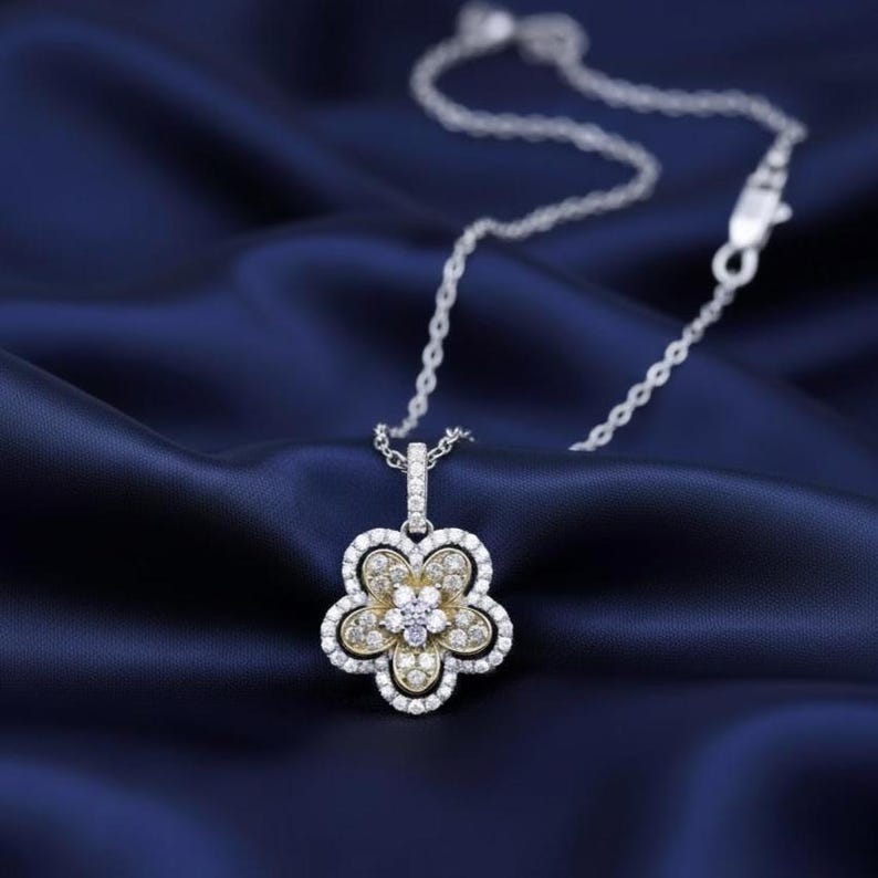 Puede incluir: Un collar de plata con un colgante de flor. El colgante tiene un centro dorado y piedras preciosas blancas alrededor de los p&eacute;talos. El collar est&aacute; sobre un fondo de sat&eacute;n azul oscuro.