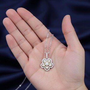Puede incluir: Un collar de plata con un colgante de flor. El colgante tiene un grupo central de piedras peque&ntilde;as y claras rodeadas por un anillo de piedras claras m&aacute;s grandes. Los p&eacute;talos exteriores est&aacute;n delineados con piedras claras. El collar se sostiene en una mano sobre un fondo azul oscuro.