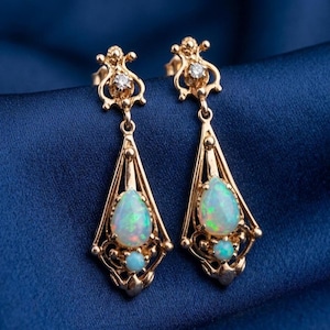 Könnte beinhalten: Ein Paar goldfarbene Ohrhänger mit Opal-Edelsteinen. Jeder Ohrring hat einen tropfenförmigen Opal, einen kleineren runden Opal und einen kleinen Diamanten. Die Ohrringe sind vor einem dunkelblauen Hintergrund platziert.
