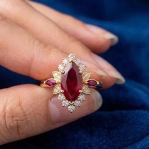 Puede incluir: Un anillo de oro con un gran rubí central en forma de marquesa, rodeado de diamantes más pequeños y detalles de rubí. El anillo tiene un diseño floral con un halo de pequeños diamantes alrededor del rubí.