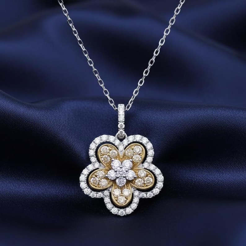 Puede incluir: Un collar con colgante en forma de flor con una cadena plateada. El colgante presenta un centro dorado rodeado de piedras preciosas blancas y doradas, con un halo de piedras preciosas blancas alrededor de los p&eacute;talos. El collar se exhibe sobre una tela azul oscuro.