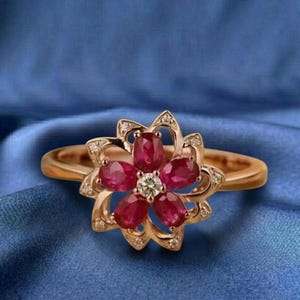 May include: A gold-toned ring featuring a flower design. The flower is composed of oval-shaped red gemstones and a central diamond. Small diamonds are set around the flower petals. The ring is set against a blue background.