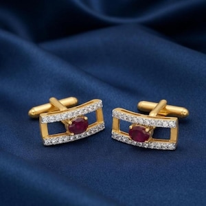May include: A pair of gold-toned rectangular cufflinks featuring a central oval-cut deep red ruby gemstone. The frame of each cufflink is accented with a row of small, clear, sparkling stones along the top and bottom edges, with a T-bar closure.