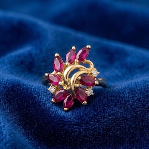 May include: A gold ring featuring a floral design with marquise-cut ruby gemstones and small diamond accents. The ring is set against a deep blue velvet background, highlighting the vibrant red of the rubies.