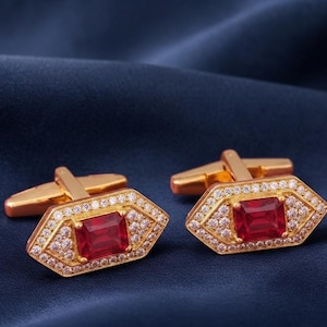 Puede incluir: Un par de gemelos dorados con una piedra roja rectangular central rodeada por un borde de pequeñas piedras transparentes. Los gemelos tienen una forma hexagonal y un mecanismo de cierre articulado en la parte posterior para la camisa.