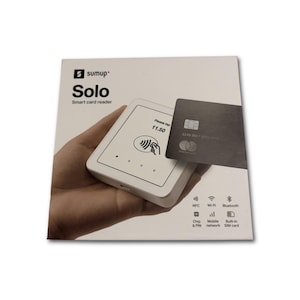 Op de afbeelding: Een witte SumUp Solo smart card reader met een zwarte creditcard. De reader toont "Please tap 11.50" en heeft NFC, Wi-Fi en Bluetooth pictogrammen. Het SumUp logo staat bovenaan.