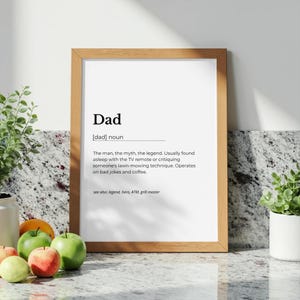 Könnte beinhalten: Ein gerahmter Druck mit der Definition von "Dad". Der Text beschreibt einen Vater als Legende, der das Rasenmähen kritisiert und schlechte Witze und Kaffee mag. Der Rahmen ist hellbraun, der Hintergrund ist weiß.