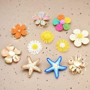 Può includere: Una collezione di abbellimenti floreali e stelle marine colorati e fatti a mano. L'assortimento comprende vari disegni floreali in tonalità arancioni, gialle, bianche e multicolore, insieme a due stelle marine, una beige e una blu. Ideale per progetti di artigianato.