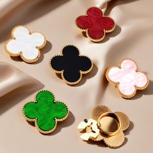 Peut inclure: Plusieurs supports de téléphone en forme de trèfle, de couleurs variées : blanc, rouge, noir, rose et vert. Chaque support a une bordure dorée et un anneau de maintien doré au dos. Présentés sur une surface satinée.