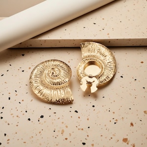 Może przedstawiać: Dwuczęściowe, złote, dekoracyjne pudełko w kształcie muszli nautilusa. Pudełko jest otwarte, ukazując małą, okrągłą komorę. Projekt muszli charakteryzuje się skomplikowanymi detalami i spiralnym wzorem. Pudełko znajduje się na nakrapianej powierzchni.