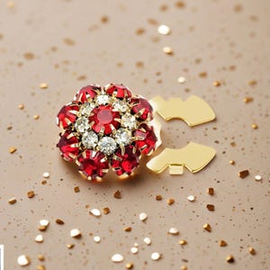 Puede incluir: Broche floral con piedras preciosas rojas y transparentes engastadas en metal dorado. El broche tiene un grupo central de piedras transparentes rodeadas de piedras rojas. Dos cierres de metal dorado están junto al broche.