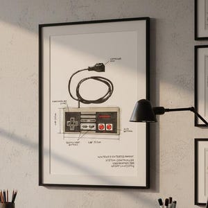 Puede incluir: Impresión enmarcada que presenta un diagrama detallado de un controlador de Nintendo Entertainment System. La ilustración incluye dimensiones, etiquetas de botones y un cable conectado. La impresión se muestra en un escritorio de madera con una lámpara y útiles de oficina.