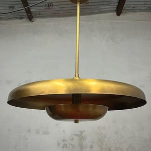 Op de afbeelding: Een messingkleurige hanglamp in een mid-century modern stijl. Het heeft een grote, platte, cirkelvormige kap en een kleinere, komvormige kap. De lamp hangt aan een lange staaf en een cirkelvormige kap.