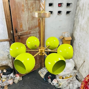Puede incluir: Una lámpara de araña moderna con seis pantallas de globo de color chartreuse. La lámpara tiene un marco de color latón y está suspendida de una cadena. La lámpara está diseñada para uso en interiores.