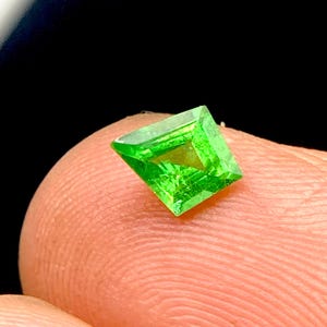 Könnte beinhalten: Ein leuchtender, quadratisch geschliffener grüner Edelstein mit einer facettierten Oberfläche, die das Licht reflektiert. Der Stein zeigt einen brillanten, gesättigten smaragdgrünen Farbton und ist so positioniert, dass seine geometrische Form und klare, transparente Kristallstruktur zur Geltung kommen.