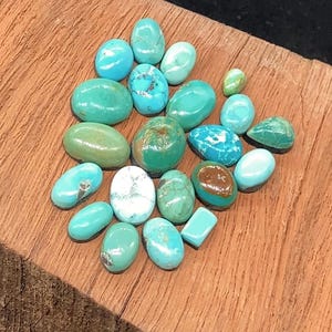 Peut inclure: Une collection de vingt et un cabochons de turquoise polis dans diverses nuances de bleu et de vert, présentant des motifs de matrice naturels, des surfaces lisses et arrondies, ainsi que des formes ovales, rondes et rectangulaires.