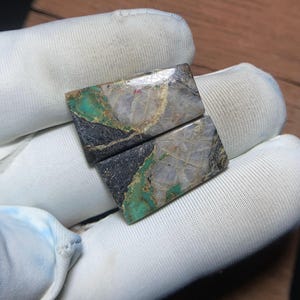 Può includere: Due cabochon rettangolari caratterizzati da un mix di quarzo bianco, venature turchesi vibranti e una matrice grigio scuro. Le pietre presentano una superficie lucida e brillante con motivi naturali distinti e una distribuzione irregolare.